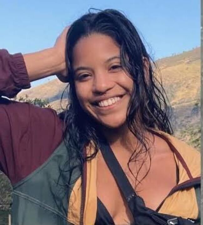 Mulher sorridente com piercing no nariz, cabelo molhado e mochila.
