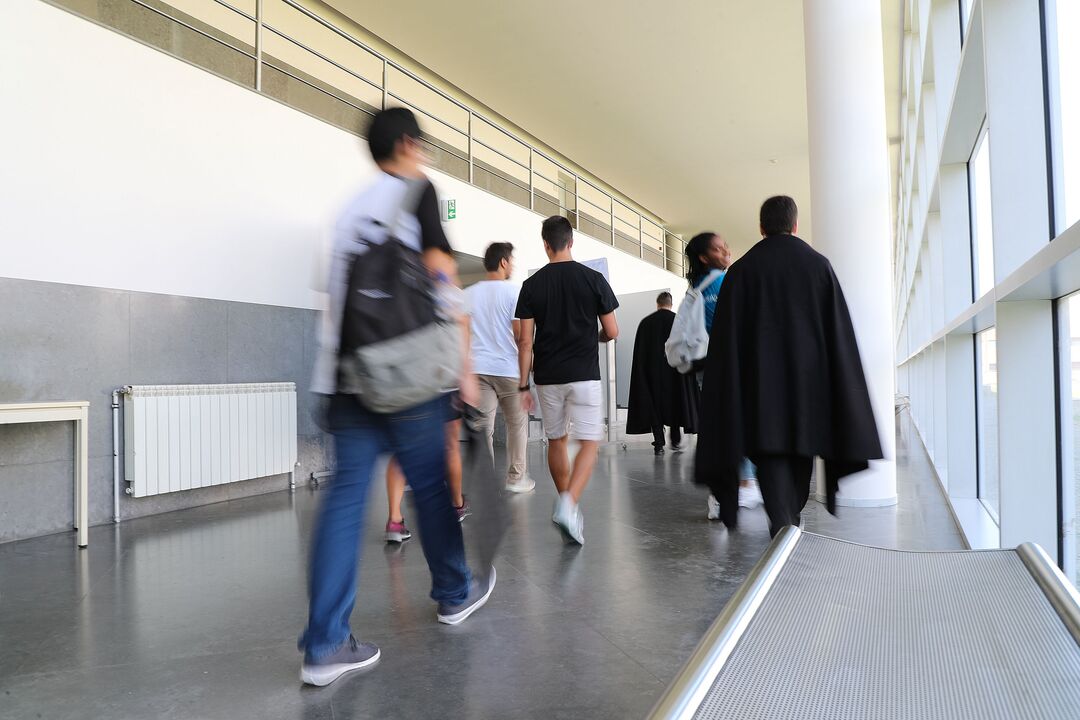 Pessoas caminham num corredor com luz natural, algumas com capas pretas