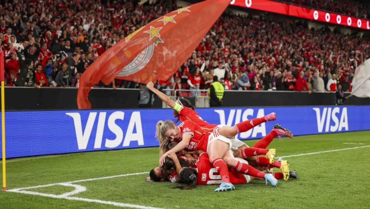 Jogadoras celebram vitória em campo, com adeptos e bandeira ao fundo