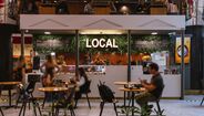 Pessoas almoçam no restaurante 'Local', com decoração de plantas e iluminação neon
