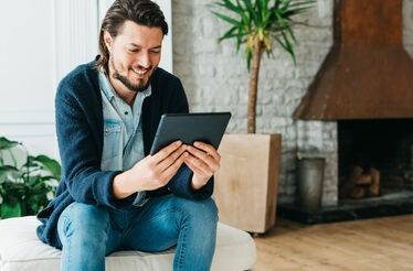 Homem sorri enquanto usa tablet, com lareira e planta no fundo.
