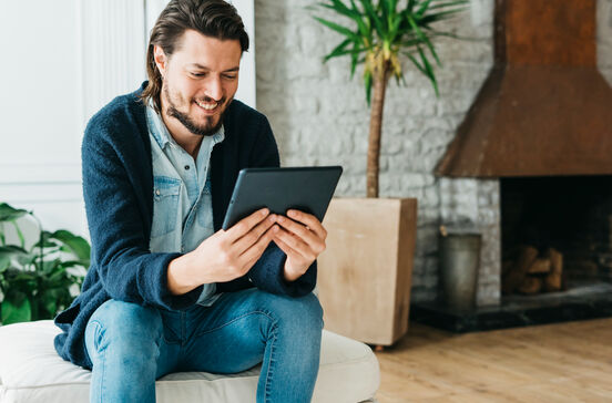 Homem sorri enquanto usa tablet, com lareira e planta no fundo.
