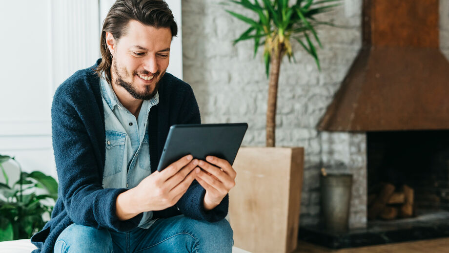 Homem sorri enquanto usa tablet, com lareira e planta no fundo.