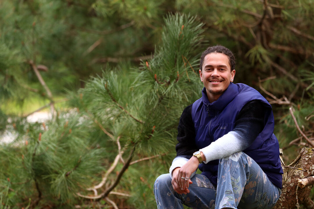 Homem sorri perto de pinheiro com colete azul e calças florais