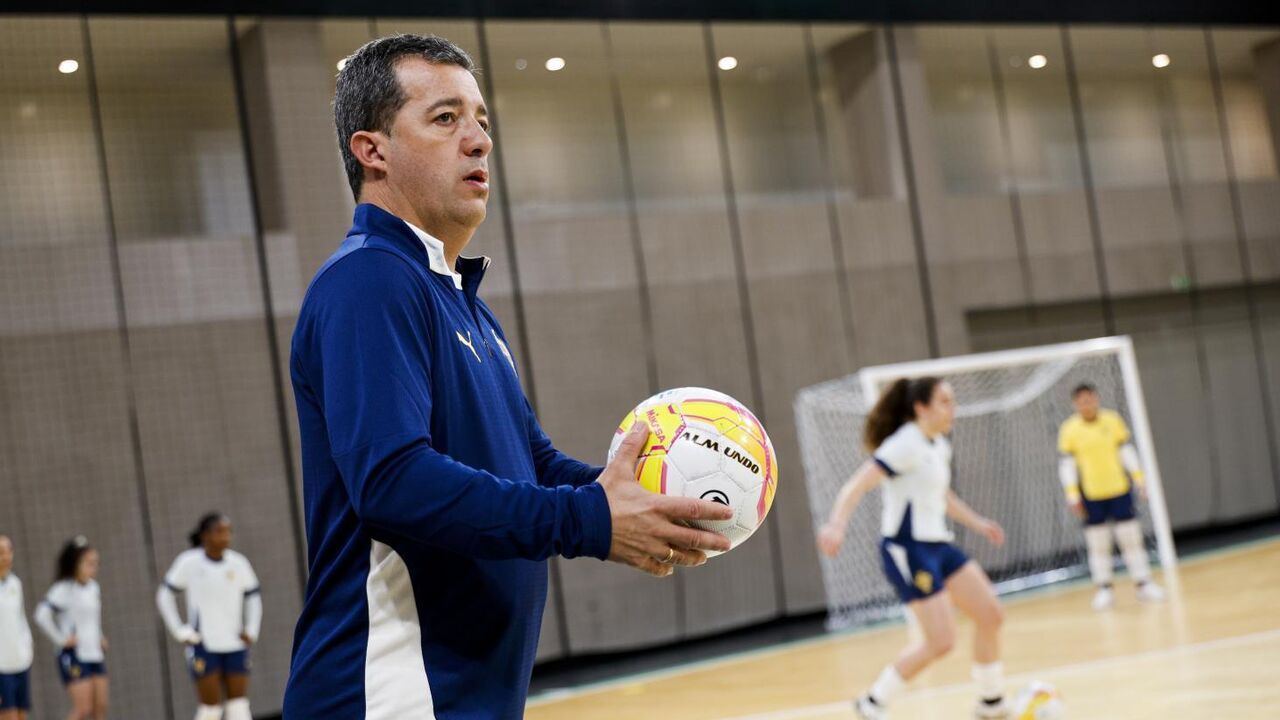 Treinador com bola observa jogadoras em treino de futsal