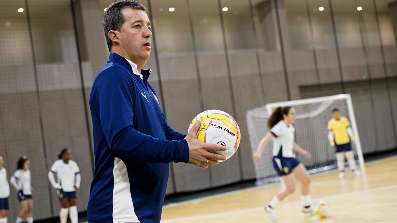 Treinador com bola observa jogadoras em treino de futsal
