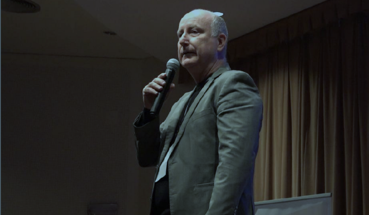 Homem discursa ao microfone num palco com cortinas