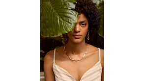 Mulher posa com colar e brincos dourados perto de uma grande folha verde