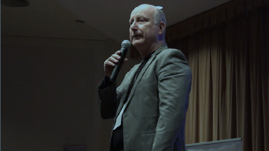 Homem discursa ao microfone num palco com cortinas