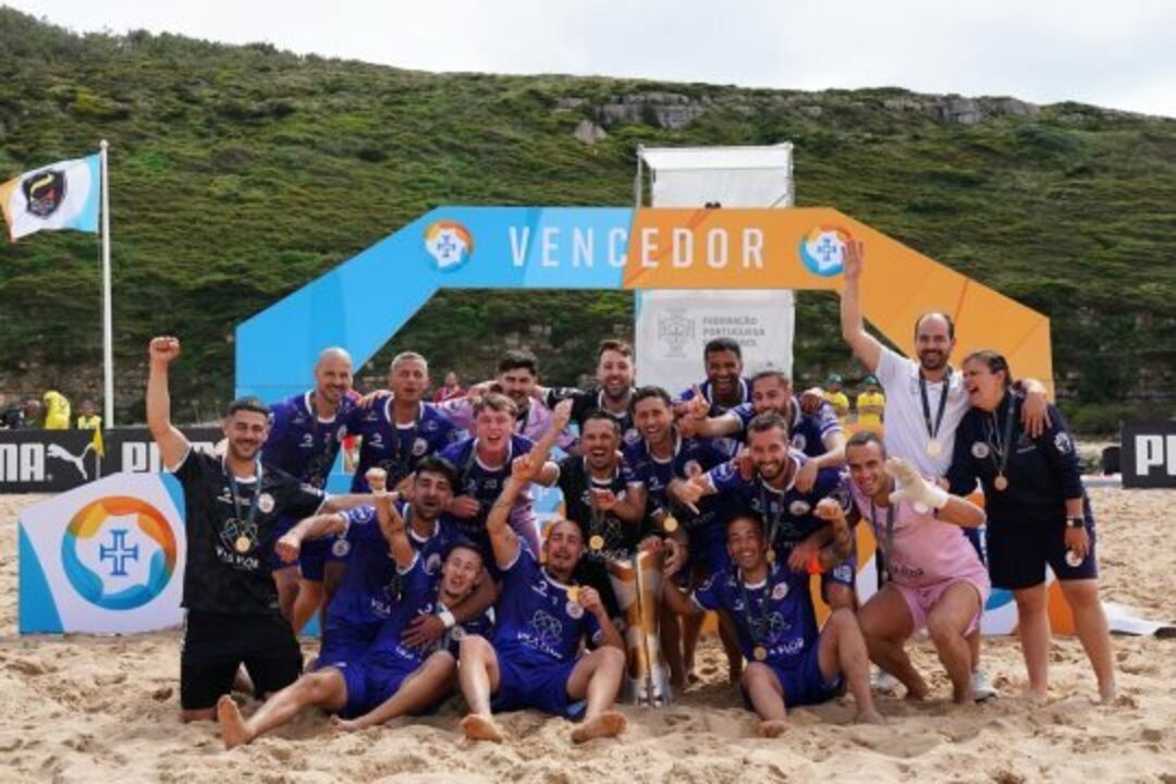 Equipa de futebol de praia celebra vitória em frente a uma faixa de "Vencedor"