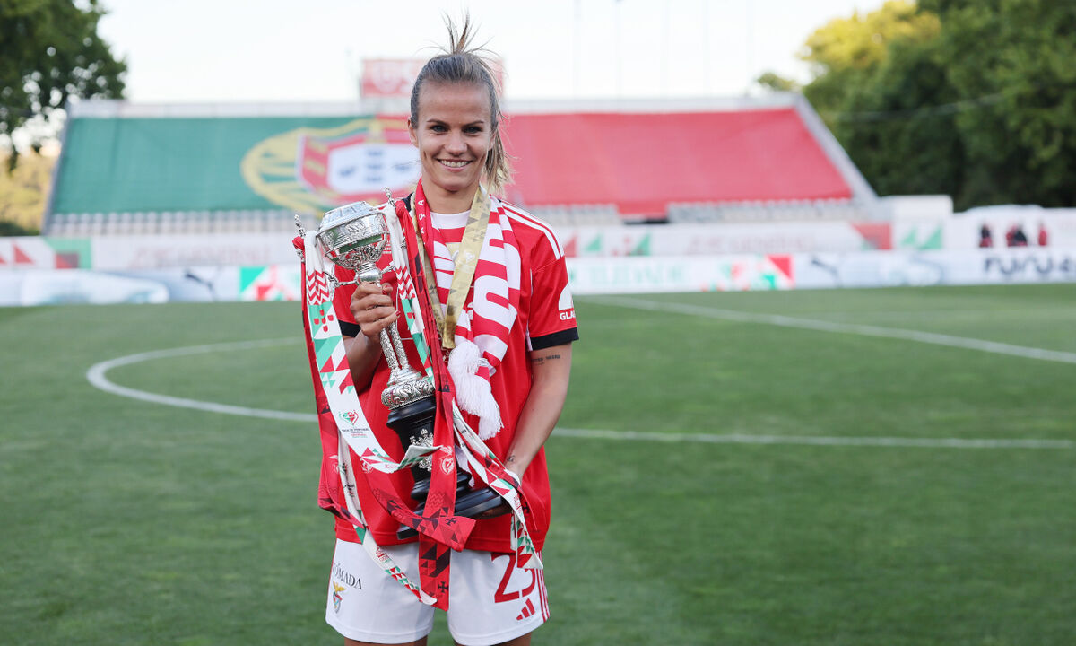 Jogadora com troféu em campo, estádio com bandeira de Portugal ao fundo