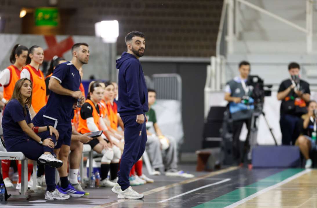 Treinador observa jogo de futsal com equipa técnica e filmagens no fundo