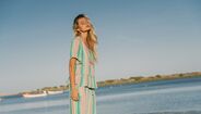 Mulher de cabelo loiro, de olhos fechados, usa um vestido às riscas coloridas numa praia
